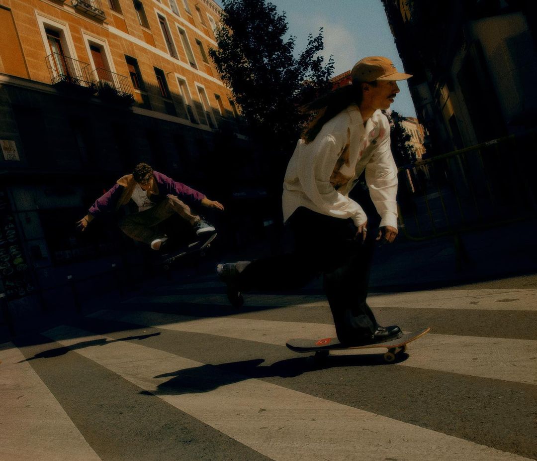 Two skateboarders perform street skating in an urban setting with buildings in the background. One skateboarder jumps while the other rides.
