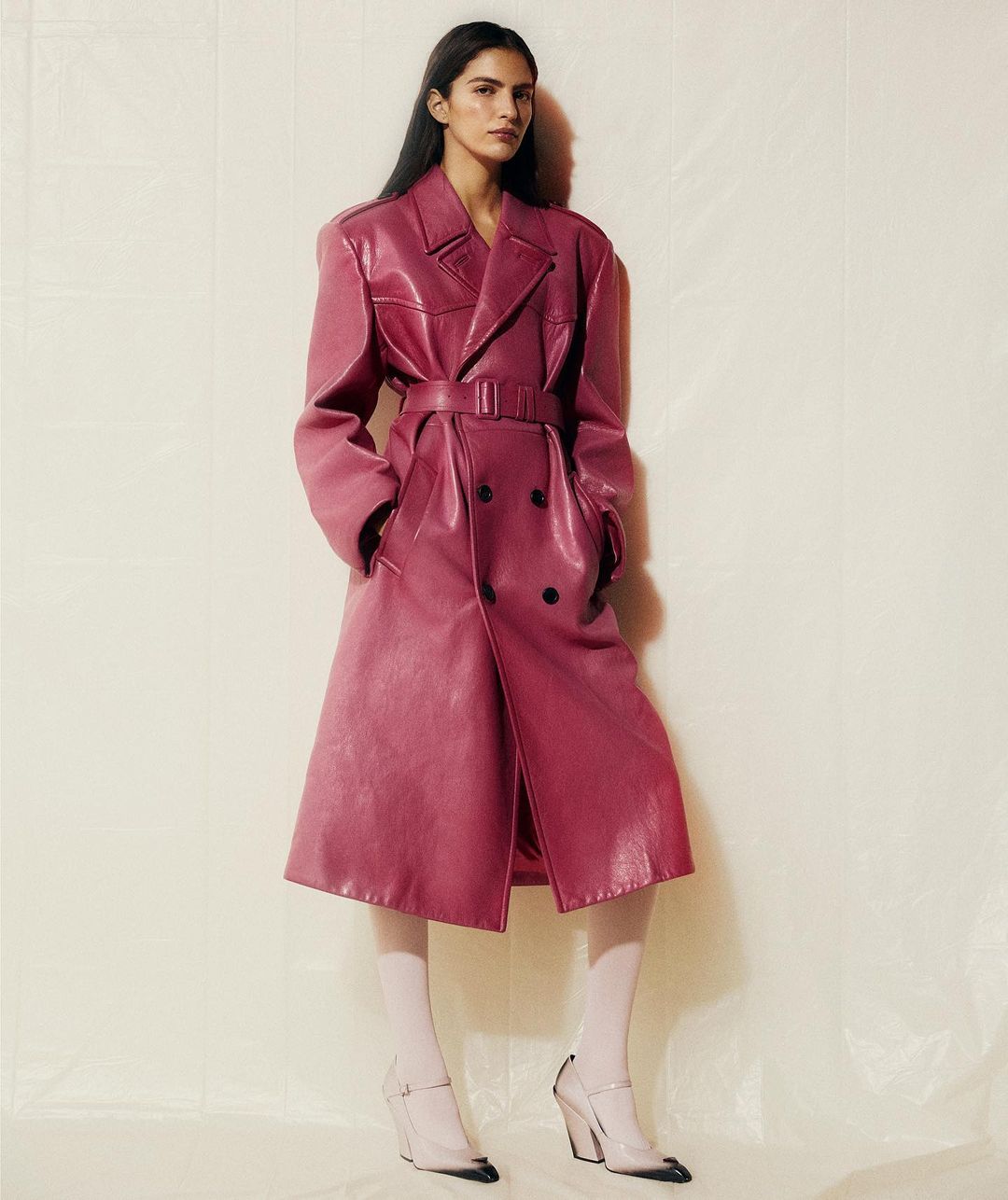 Fashionable woman in a long magenta double-breasted faux leather trench coat and white tights poses in a neutral studio environment.