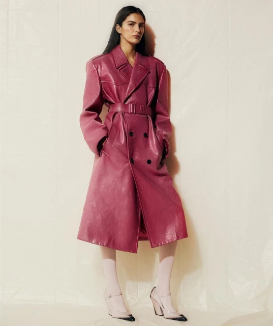 Fashionable woman in a long magenta double-breasted faux leather trench coat and white tights poses in a neutral studio environment.