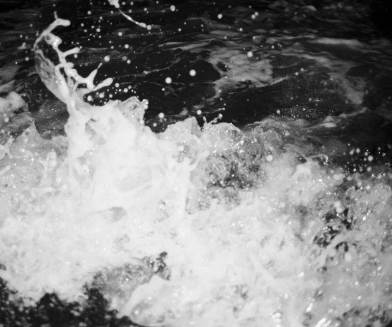 Monochromatic image showcases white water crashing and splashing against dark rocks, creating a dynamic and textured aquatic scene.