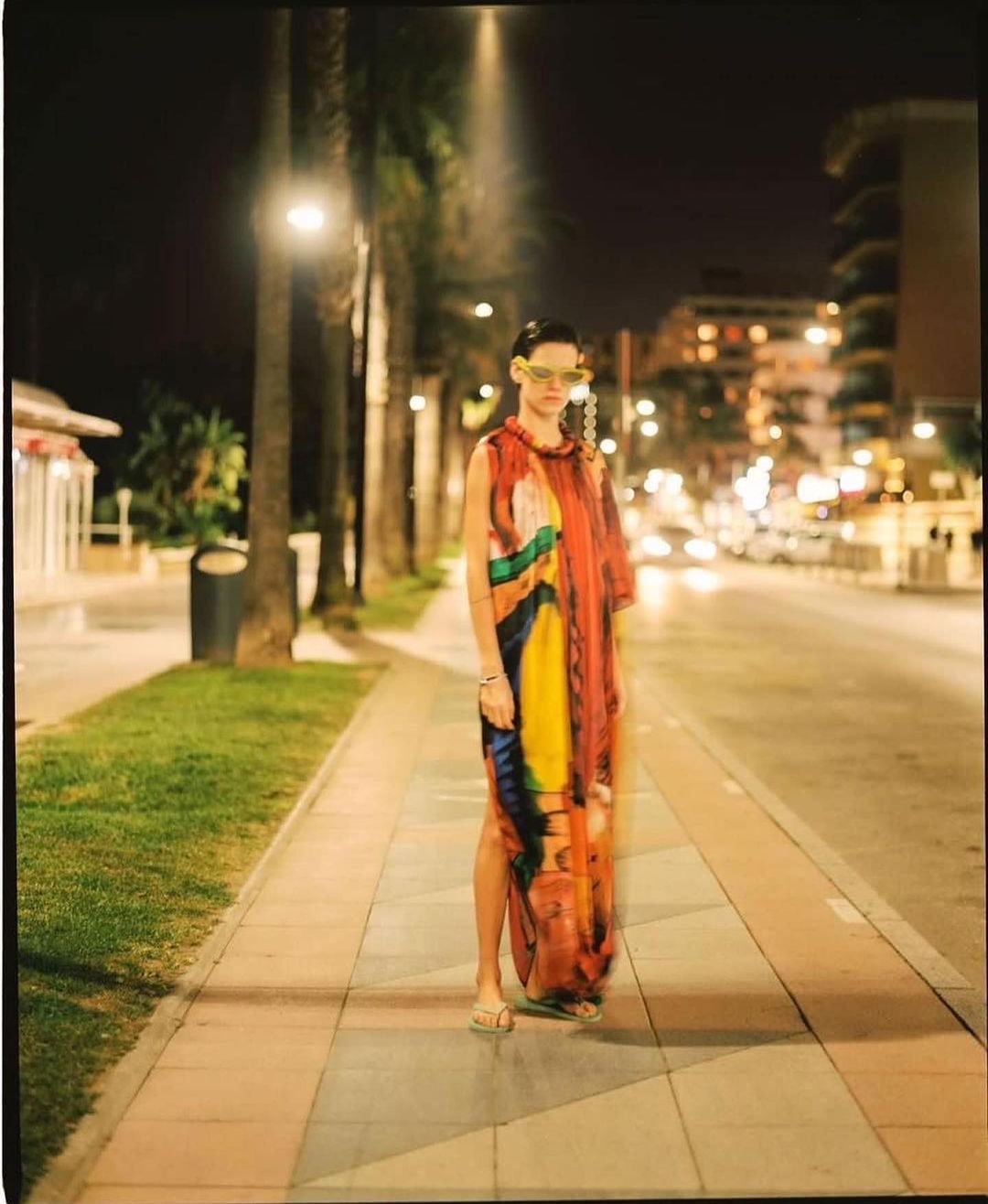 Woman in a colorful dress walking on a sidewalk at night near palm trees and streetlights, with blurred motion and an urban backdrop.