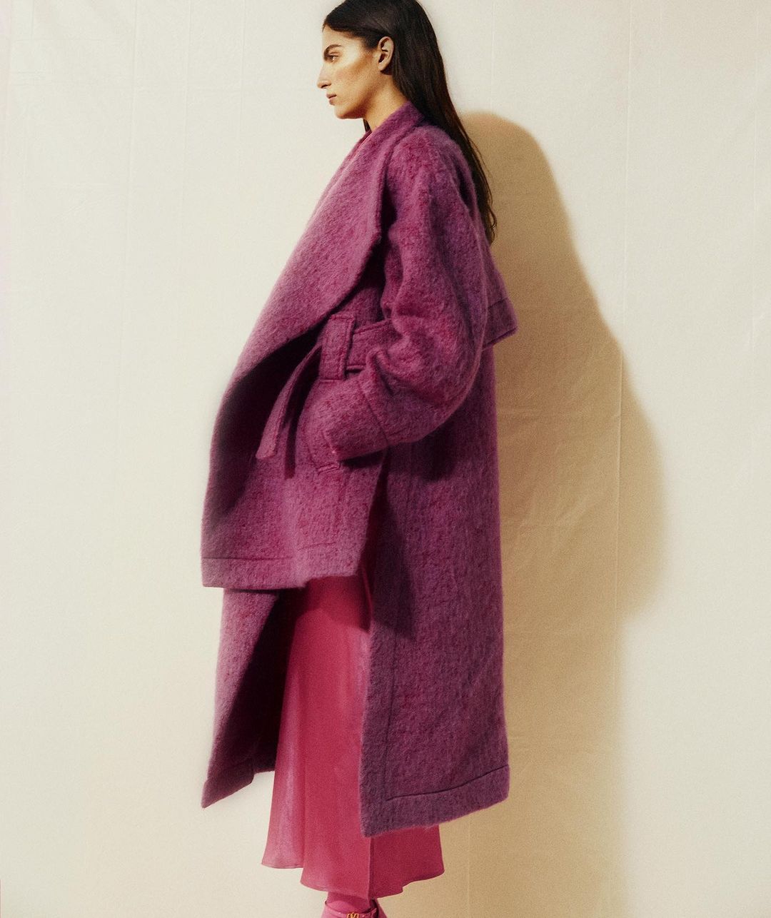 Profile shot of an adult female wearing a pink long-sleeved dress and long purple coat, posing in front of a neutral background.