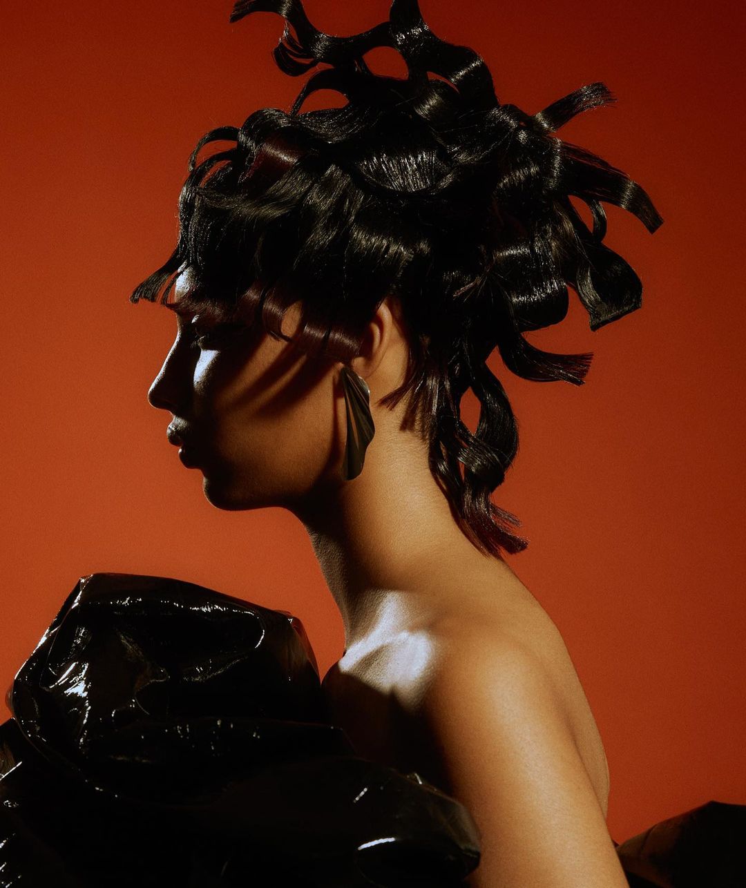 Profile shot of an adult female with unique black hairstyle against a warm orange backdrop, wearing large, dark leaf-shaped earrings.