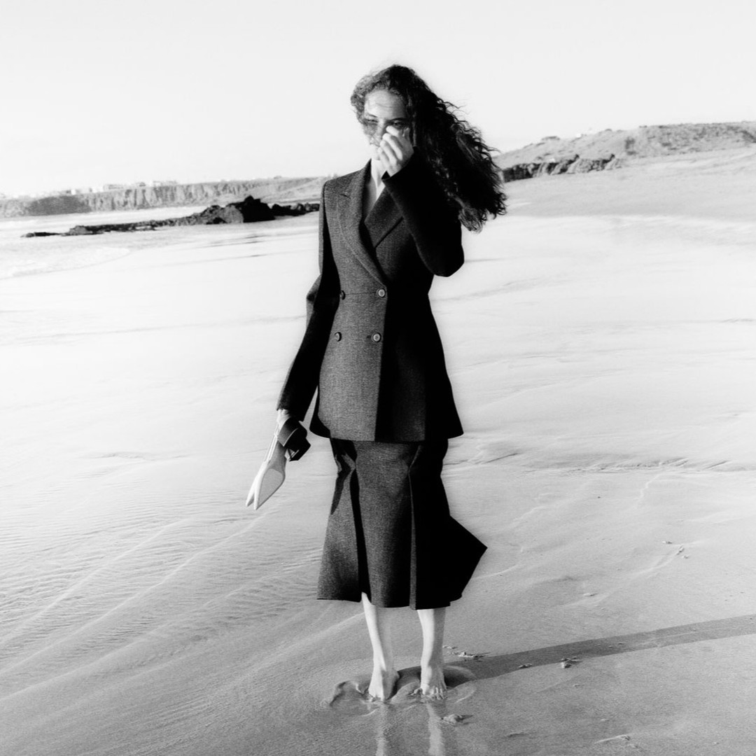 Fashion model in a black blazer and skirt walking barefoot on a beach, holding heels, with a distant shoreline in monochrome.