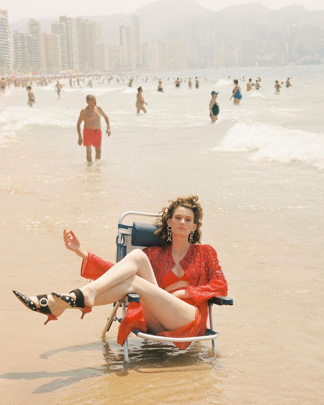 A woman in a red outfit sits in a beach chair on a sunny beach with people and buildings in the background.