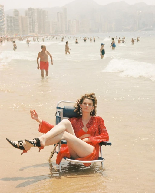 A woman in a red outfit sits in a beach chair on a sunny beach with people and buildings in the background.
