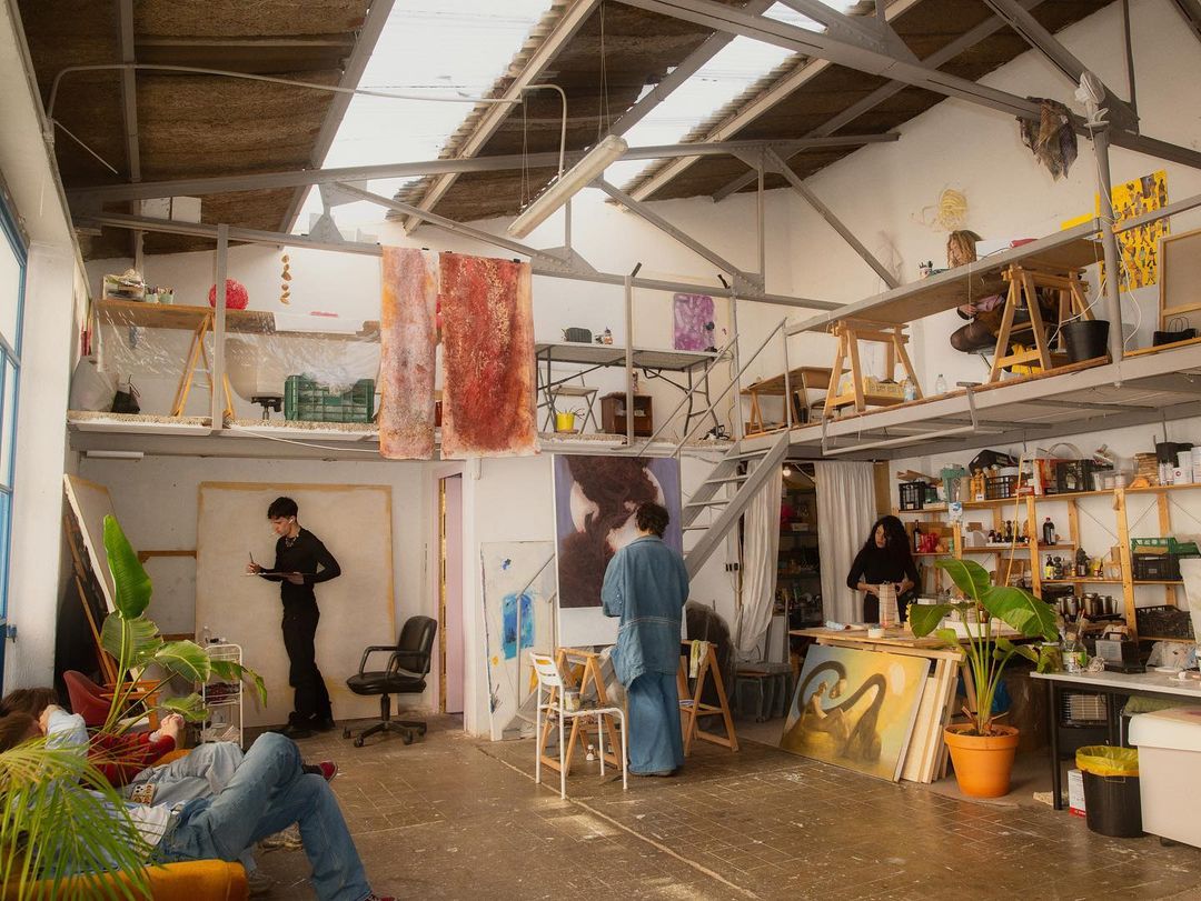 Bright and airy art studio scene with artists, paintings, plants, and loft area filled with supplies and work spaces.