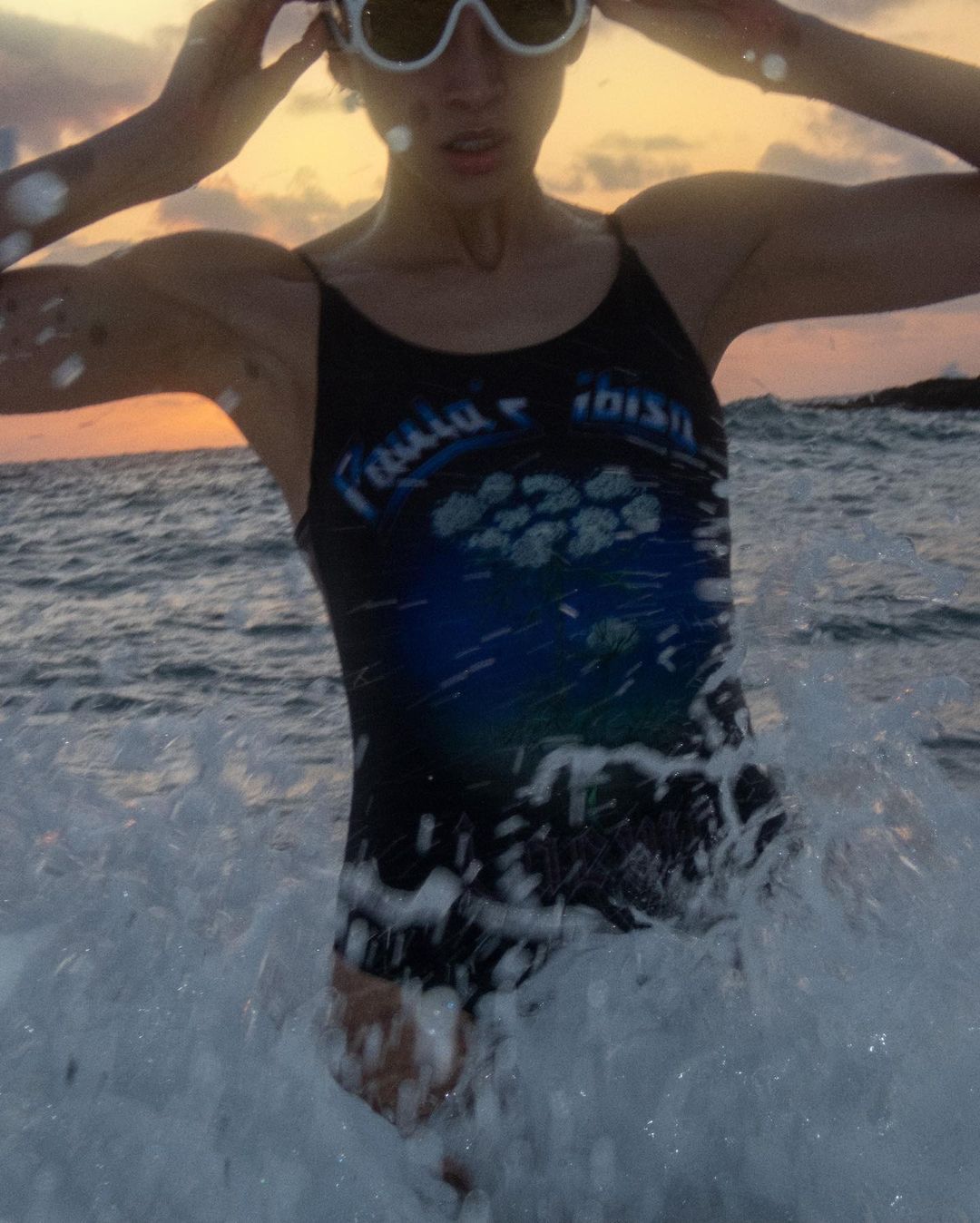 Woman in swimsuit with sunglasses emerges from the waves at the beach at sunset, creating a dynamic, summery, and active scene.
