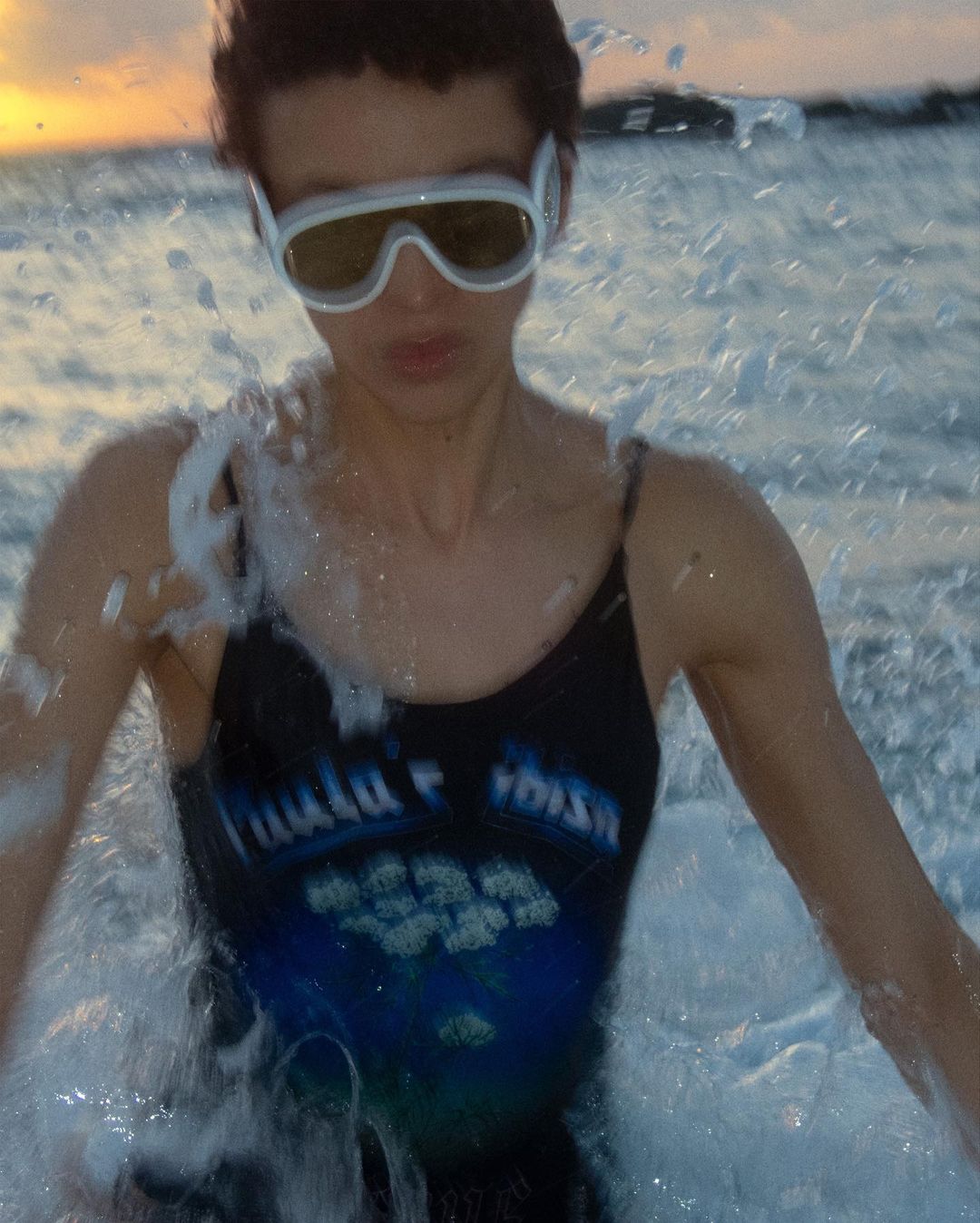 A young adult woman stands in ocean water wearing goggles and a swimsuit, with water splashing around her against a sunset sky.