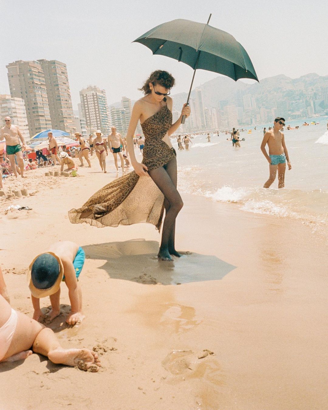 A woman in a one-shoulder swimsuit walks on a crowded beach with an umbrella. People are swimming, sunbathing, and playing in the background.