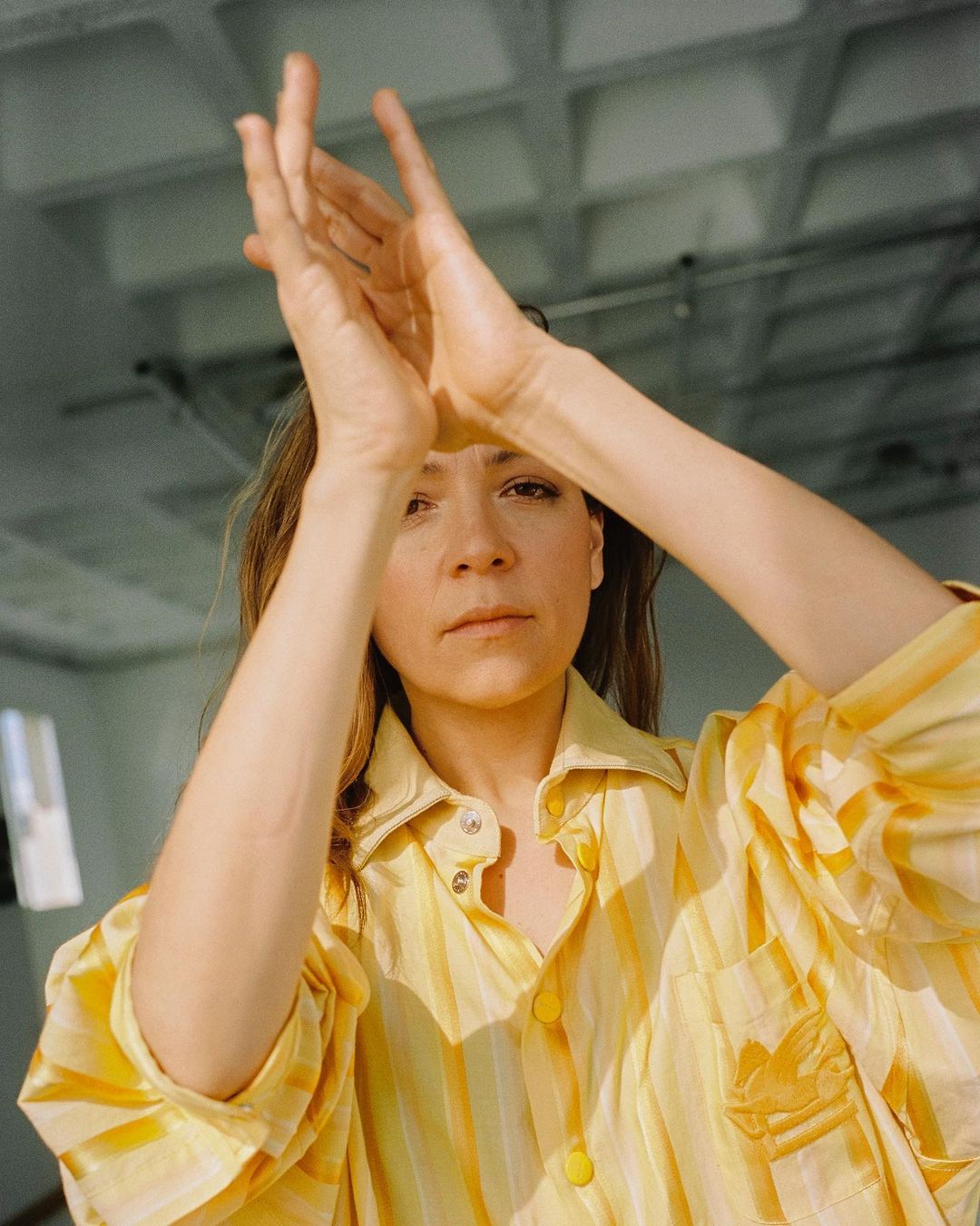 Portrait of a woman in a yellow striped shirt raising her hands in an indoor setting with sunlight creating shadows on her face and clothing.