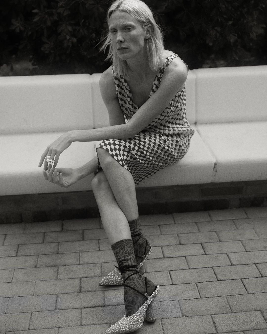 Model wearing a patterned checkered dress and embellished pointed heels sitting outdoors on a white bench directed by Pablo Curto
