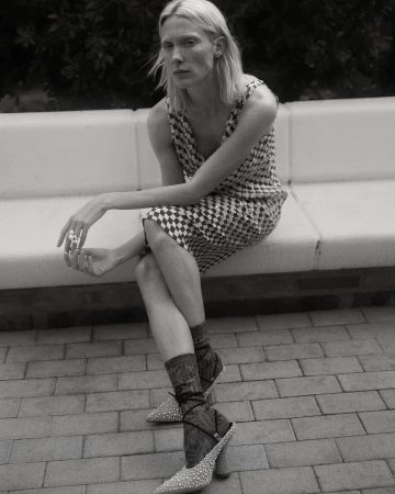 Model wearing a patterned checkered dress and embellished pointed heels sitting outdoors on a white bench directed by Pablo Curto