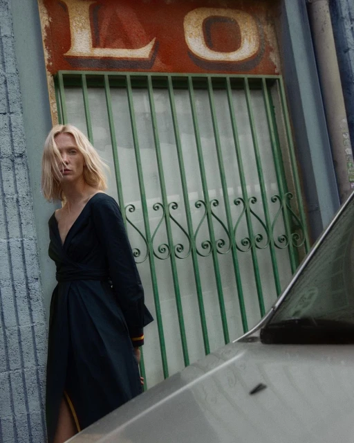 A blond woman in a dark wrap dress stands next to an old car. The scene is set against a rustic building with barred windows and worn signage.