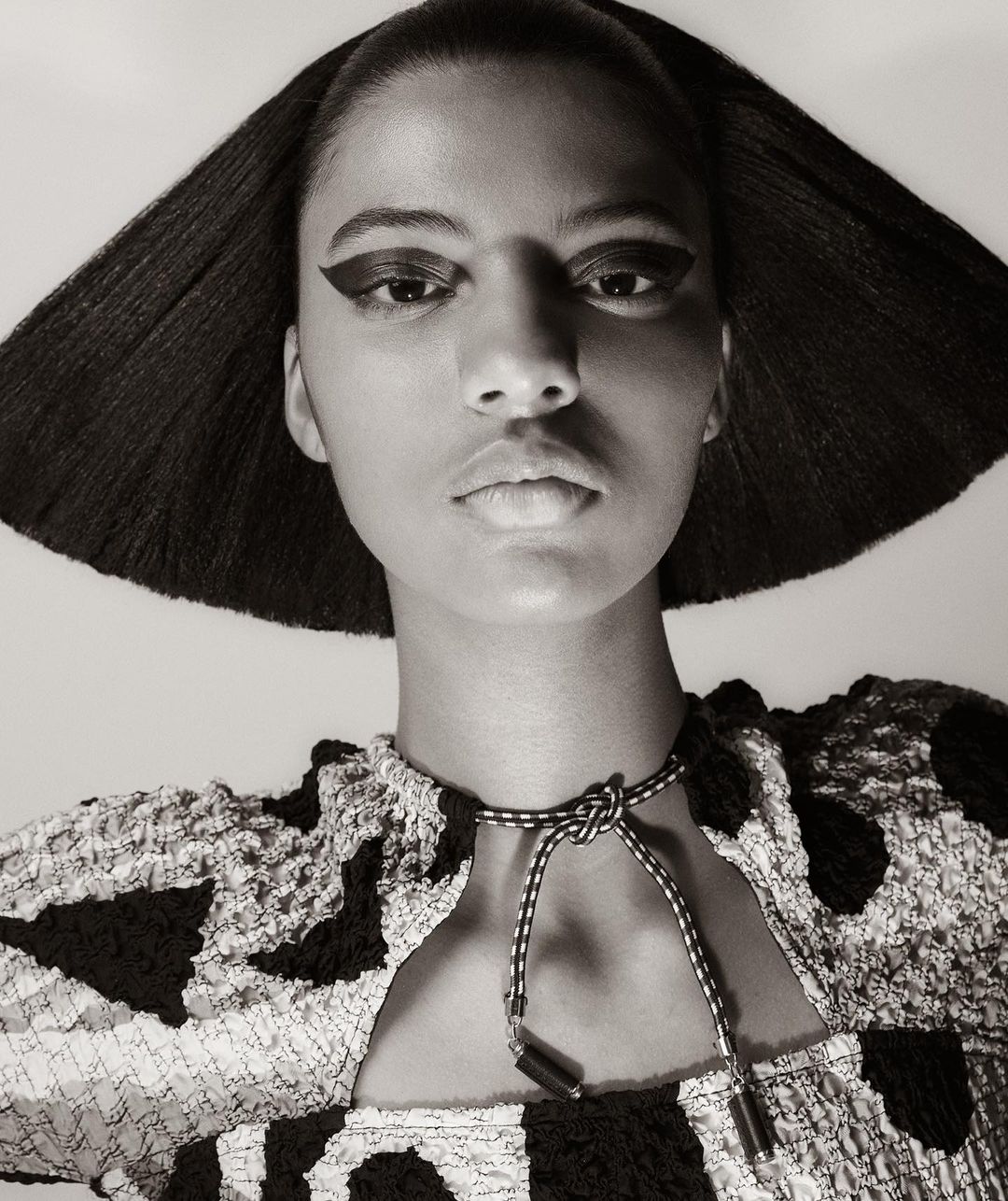 Monochrome portrait of a woman with graphic eye makeup and a patterned top and necklace, set against a neutral backdrop.
