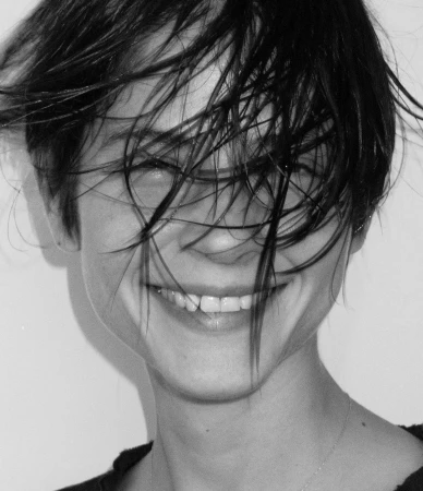 Close-up portrait of a smiling woman with dark wet hair across her face against a plain light background.