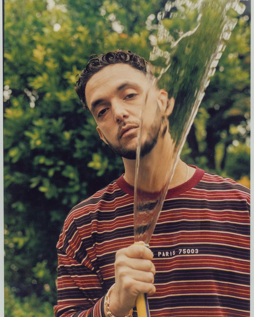 Portrait of a young adult man holding a hose with water spraying, wearing a striped shirt, outdoors with green foliage in the background.