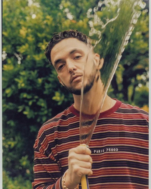 Portrait of a young adult man holding a hose with water spraying, wearing a striped shirt, outdoors with green foliage in the background.