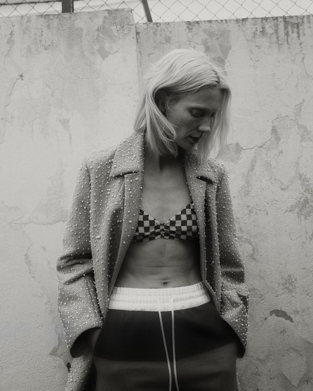 Monochrome shot of a blonde woman in a bikini top, jacket, and sweatpants posing in front of a textured wall and chain link fence.