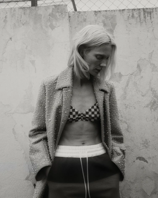 Monochrome shot of a blonde woman in a bikini top, jacket, and sweatpants posing in front of a textured wall and chain link fence.