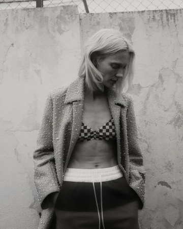 Monochrome shot of a blonde woman in a bikini top, jacket, and sweatpants posing in front of a textured wall and chain link fence.