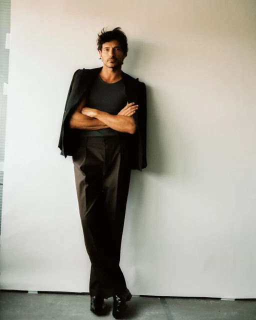 A man in a grey shirt and dark pants stands in front of a white wall wearing a blazer slung over his shoulder indoors.