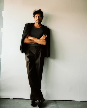 A man in a grey shirt and dark pants stands in front of a white wall wearing a blazer slung over his shoulder indoors.