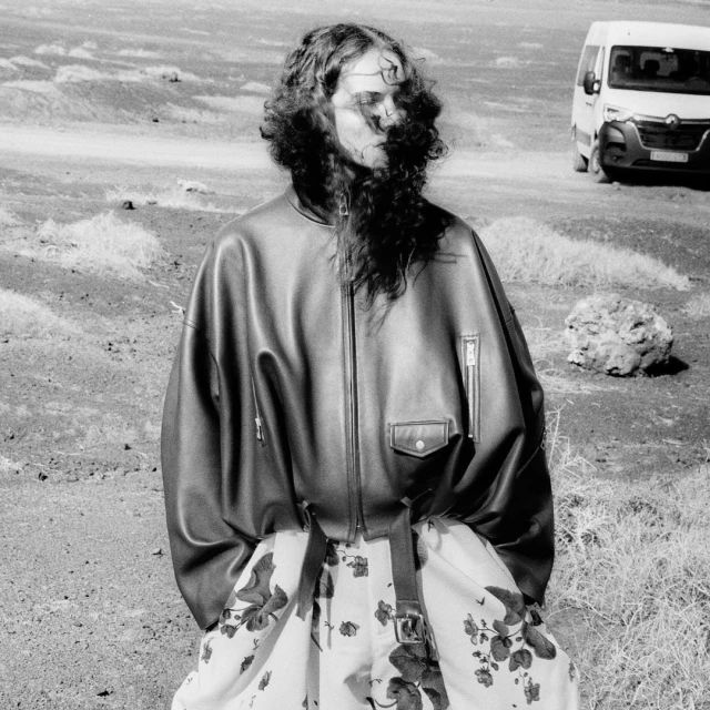 Monochrome portrait of a person in a leather bomber jacket standing in a sparse landscape with a vehicle in the background.