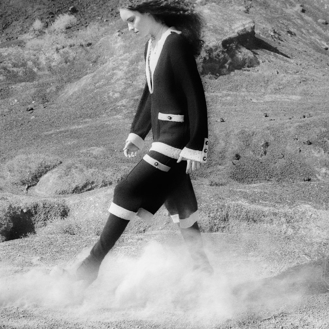 Black and white fashion shot of a woman in a jacket and shorts walking through a dusty landscape with hills in the background.