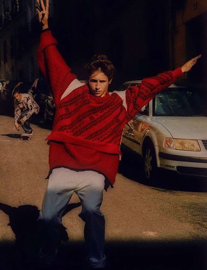 Fashionable young man in red sweater strikes dynamic pose on city street, while skateboarder rides by a parked car in the background.
