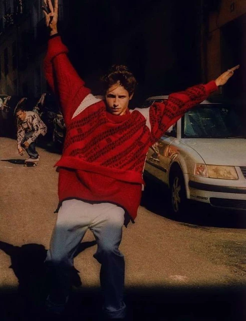 Fashionable young man in red sweater strikes dynamic pose on city street, while skateboarder rides by a parked car in the background.