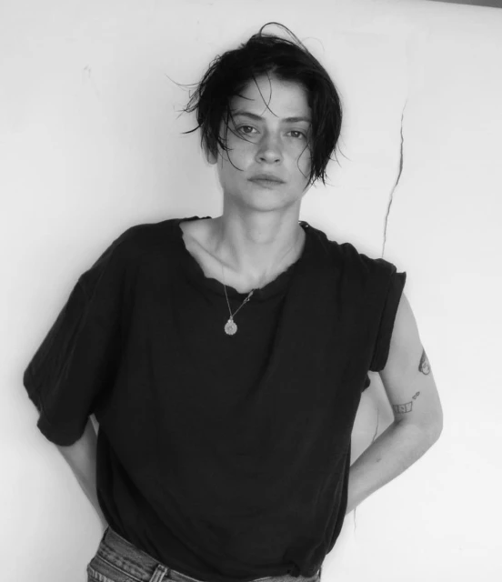 Black and white portrait of a young adult woman with short dark hair, wearing a black t-shirt and jeans, standing indoors against a white wall.