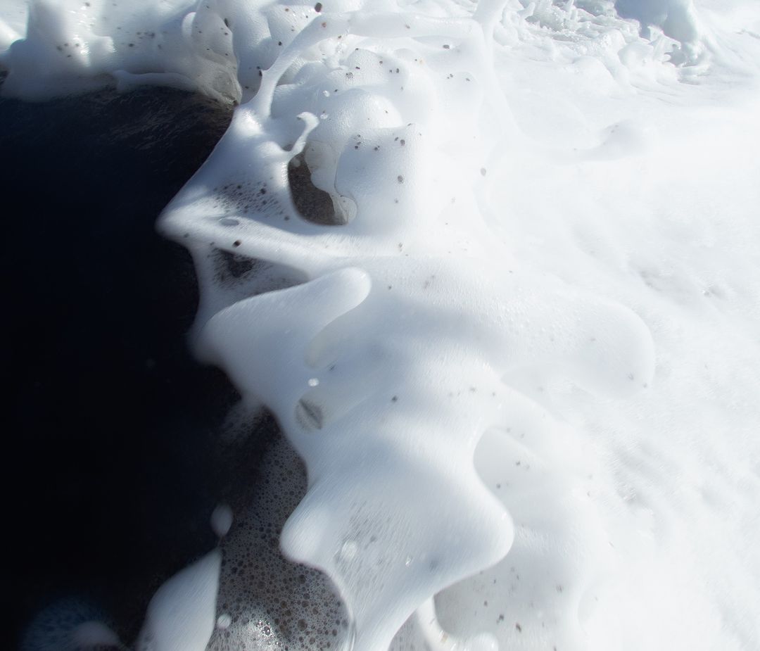 Close-up of white sea foam on a black sand beach, creating an abstract and textured coastal scene with a monochromatic color palette.