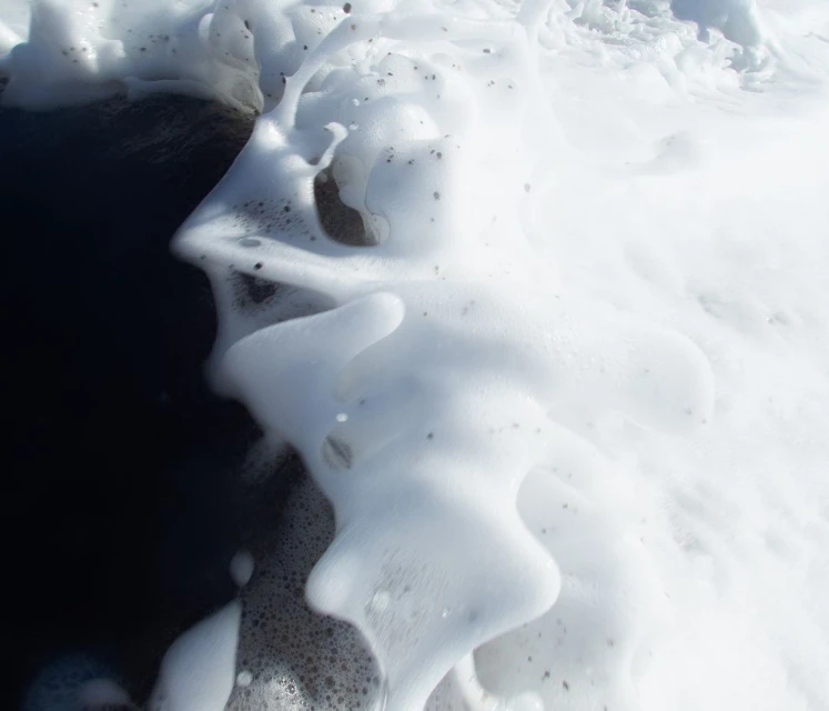 Close-up of white sea foam on a black sand beach, creating an abstract and textured coastal scene with a monochromatic color palette.