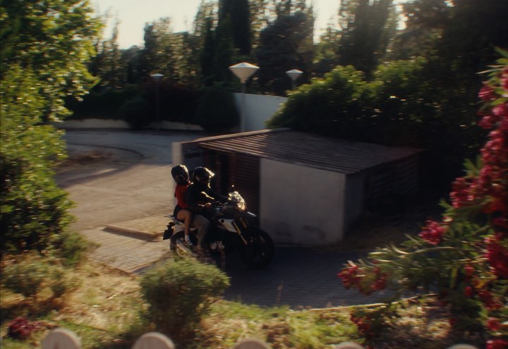 Two people riding a motorcycle on a paved road in a residential area, surrounded by greenery and buildings on a sunny day.