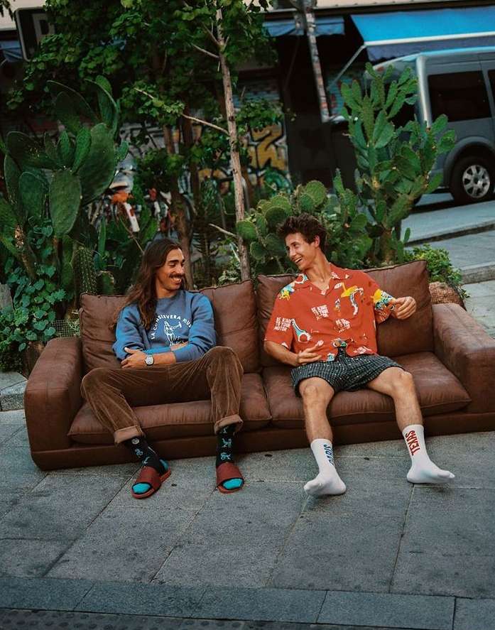 Two adult men sit on a brown couch outside amidst cacti, trees, and a parked gray van with an urban background in daylight.