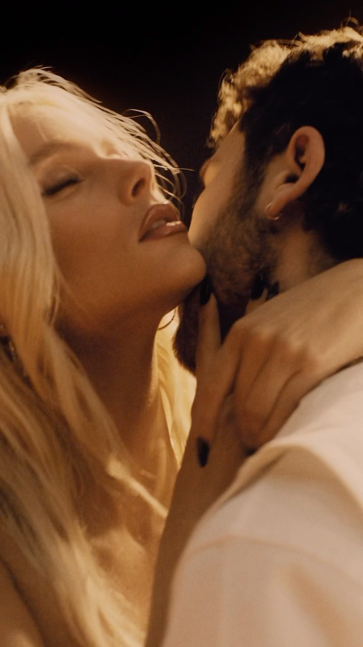 Close-up of a blonde woman with closed eyes, embraced by a man with dark, curly hair, bathed in golden light, suggesting intimacy.