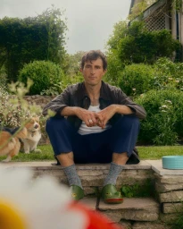 Man sitting on stone steps in a lush garden wearing a dark jacket, blue trousers, and green frog clogs, with a small dog beside him.