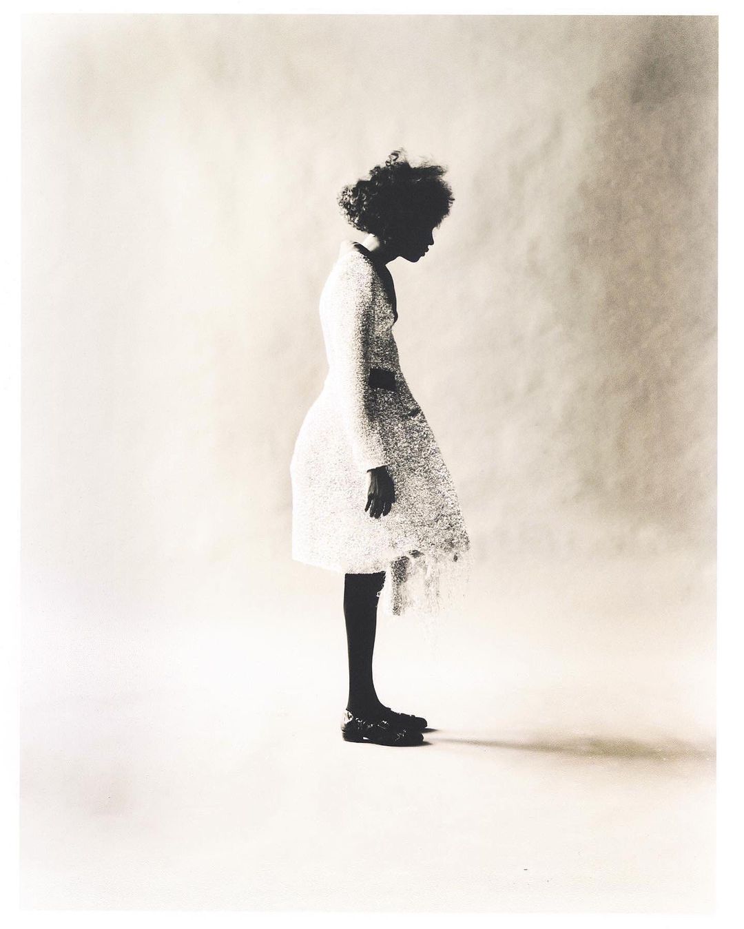 Young Black girl in a white textured dress and black shoes poses in a side profile against a soft white background in a fashion-forward photo.