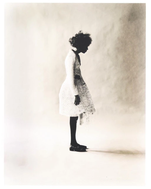 Young Black girl in a white textured dress and black shoes poses in a side profile against a soft white background in a fashion-forward photo.