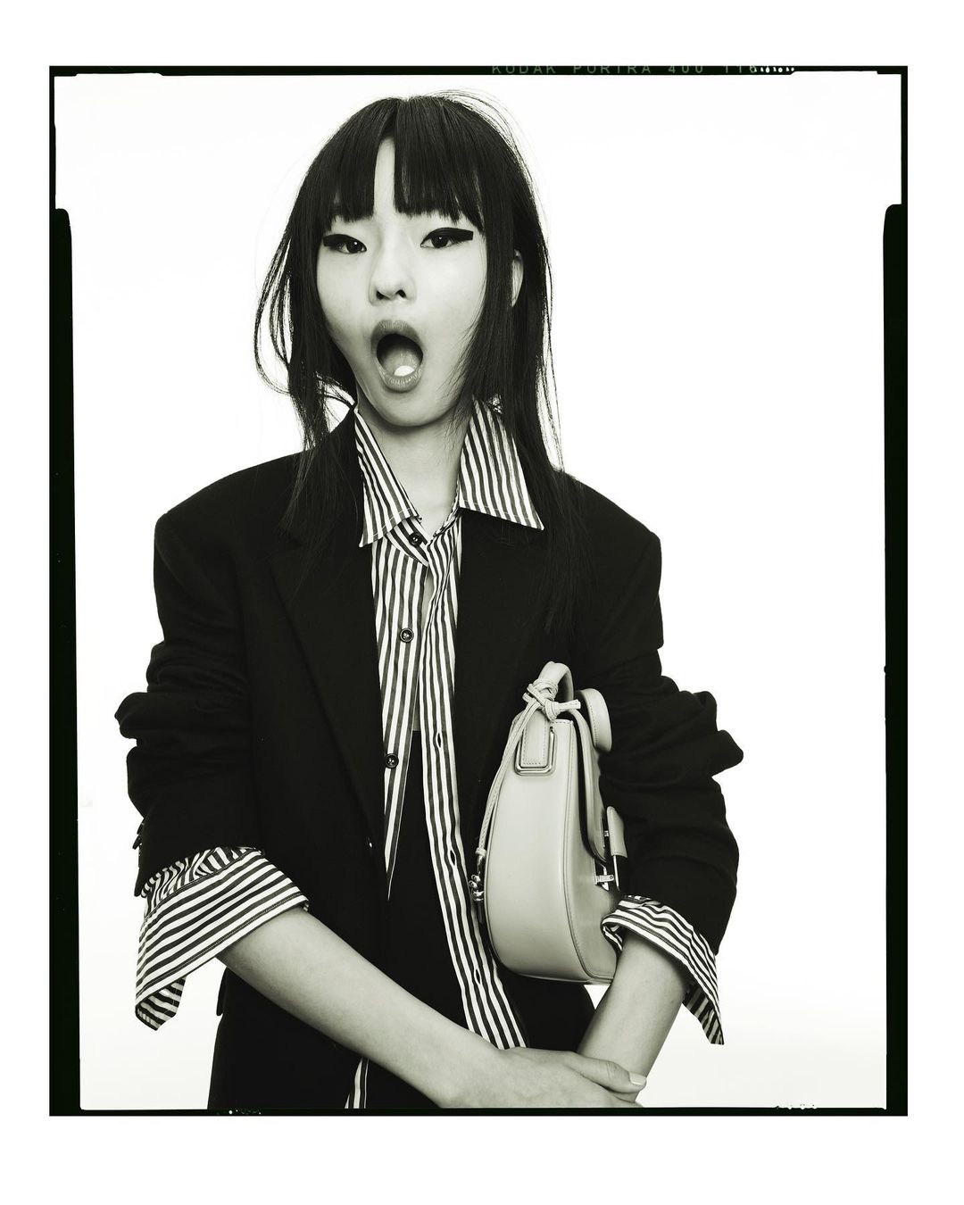 An Asian woman in a striped shirt and black blazer holds a purse against a bright background in a medium shot fashion portrait.