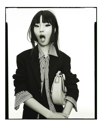 An Asian woman in a striped shirt and black blazer holds a purse against a bright background in a medium shot fashion portrait.
