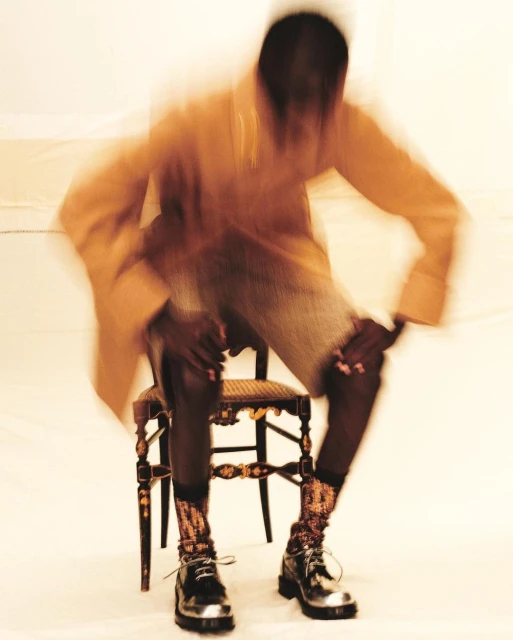 Blurred motion shot of a person with dark skin sitting on an ornate chair wearing shiny metallic boots and tan clothing in a photo studio.