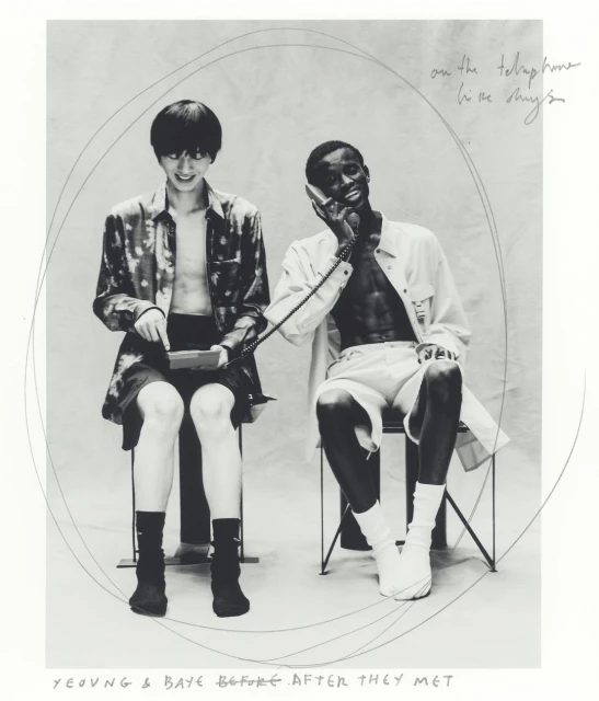 Black and white photo of two young adults seated indoors, one using a telephone while the other interacts with a handheld device.