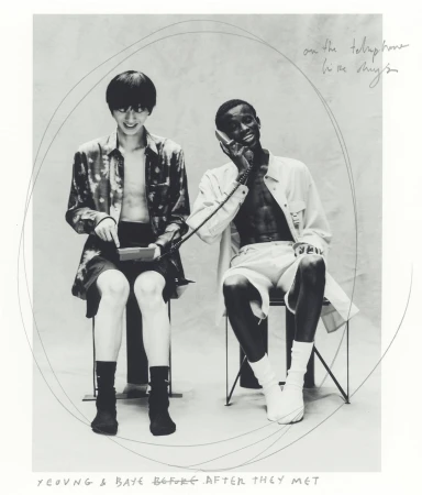 Black and white photo of two young adults seated indoors, one using a telephone while the other interacts with a handheld device.