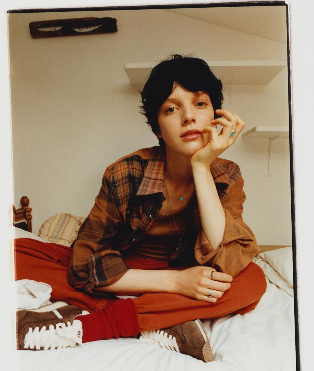 Portrait of a young adult woman with dark short hair sitting on a bed indoors, wearing a plaid shirt and orange pants, looking at the camera.