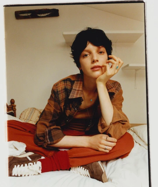 Portrait of a young adult woman with dark short hair sitting on a bed indoors, wearing a plaid shirt and orange pants, looking at the camera.