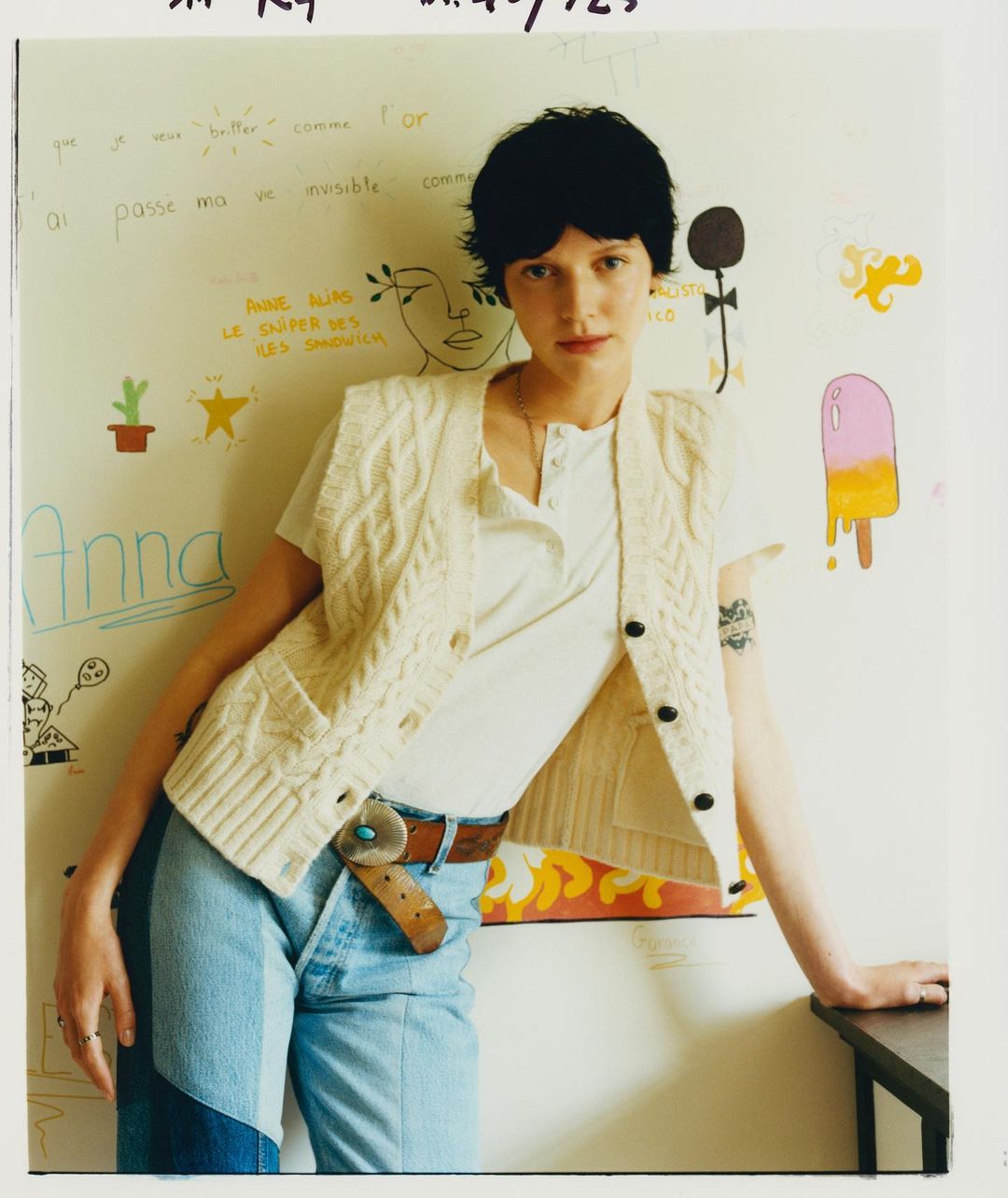 Young adult posing indoors against a wall decorated with hand-drawn art, wearing a cable-knit vest and jeans, casual fashion.