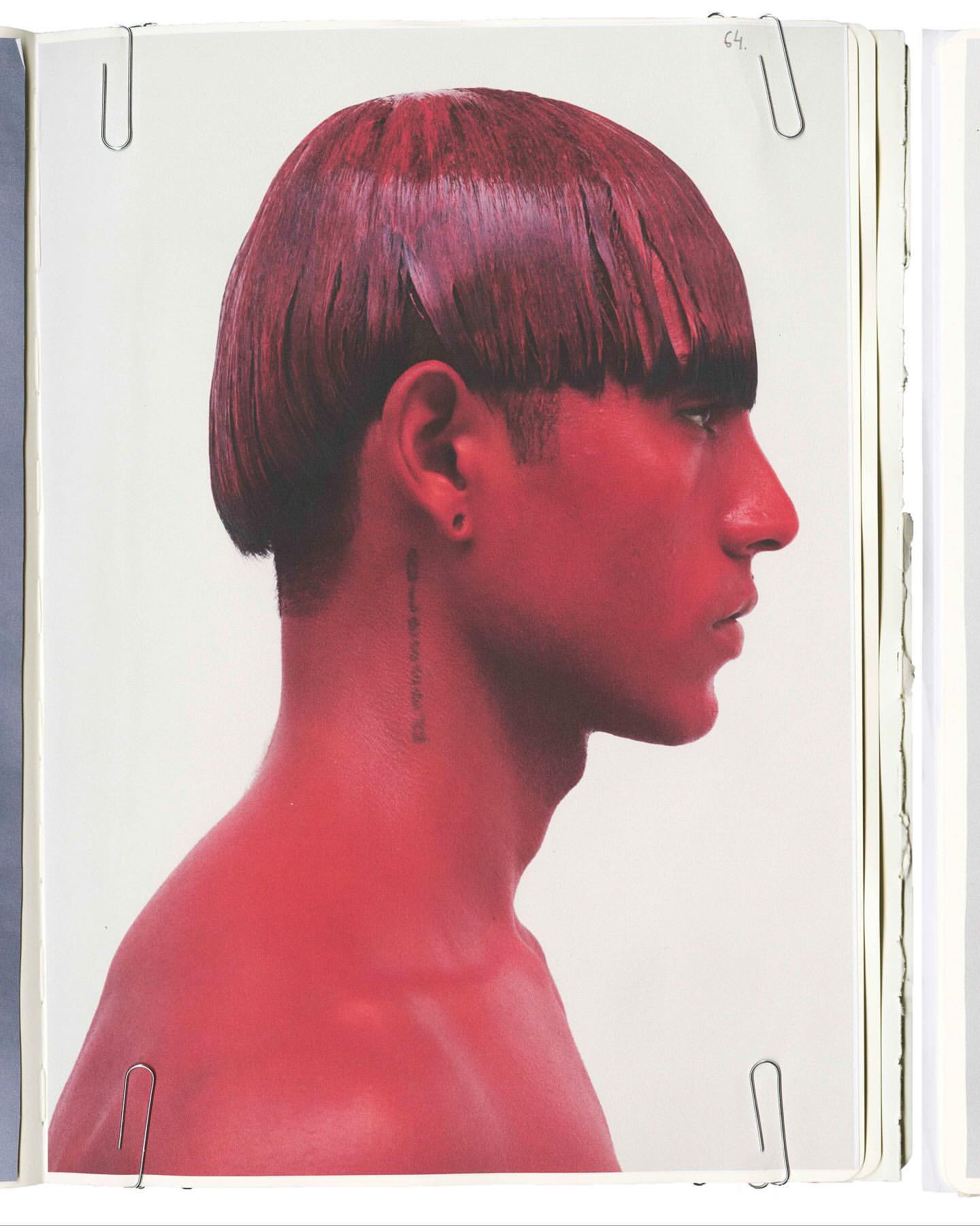 Profile of a man with red-tinted skin and dark red hair, styled with short, blunt bangs, standing against a bright white backdrop.