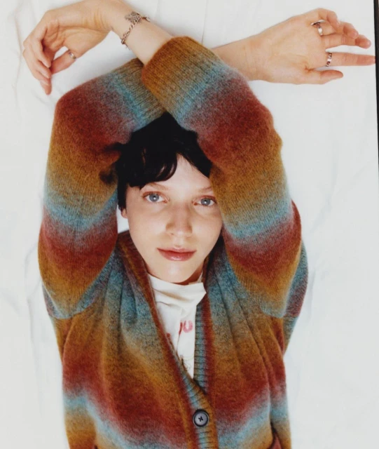 Portrait of a young woman with short brown hair wearing a striped colorful cardigan, posing with arms crossed above her head indoors.